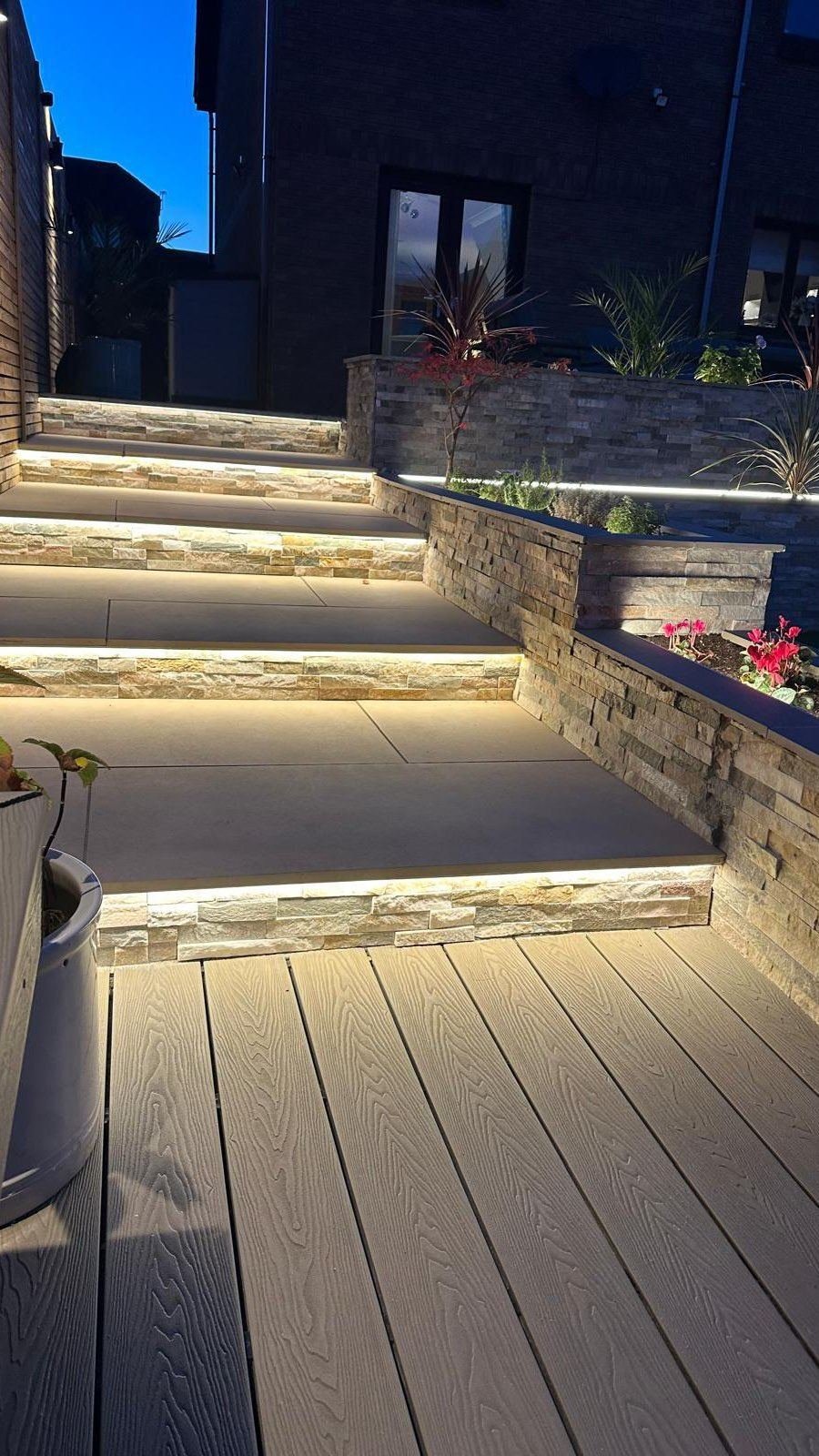 Steps down a sloped garden, each lit along the edge, beside a wall of stacked stone cladding.