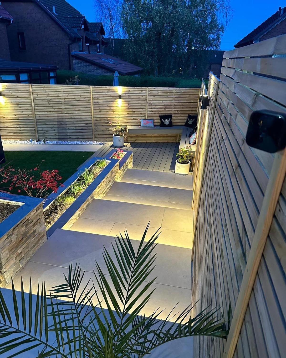 Lit porcelain steps between a stone-clad planter and slat fencing, leading down to a seating area under wall lights.