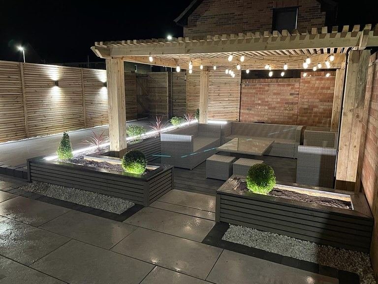 A timber pergola strung with festoon lights over outdoor sofas, with lit planters and wall lights on slat fencing.