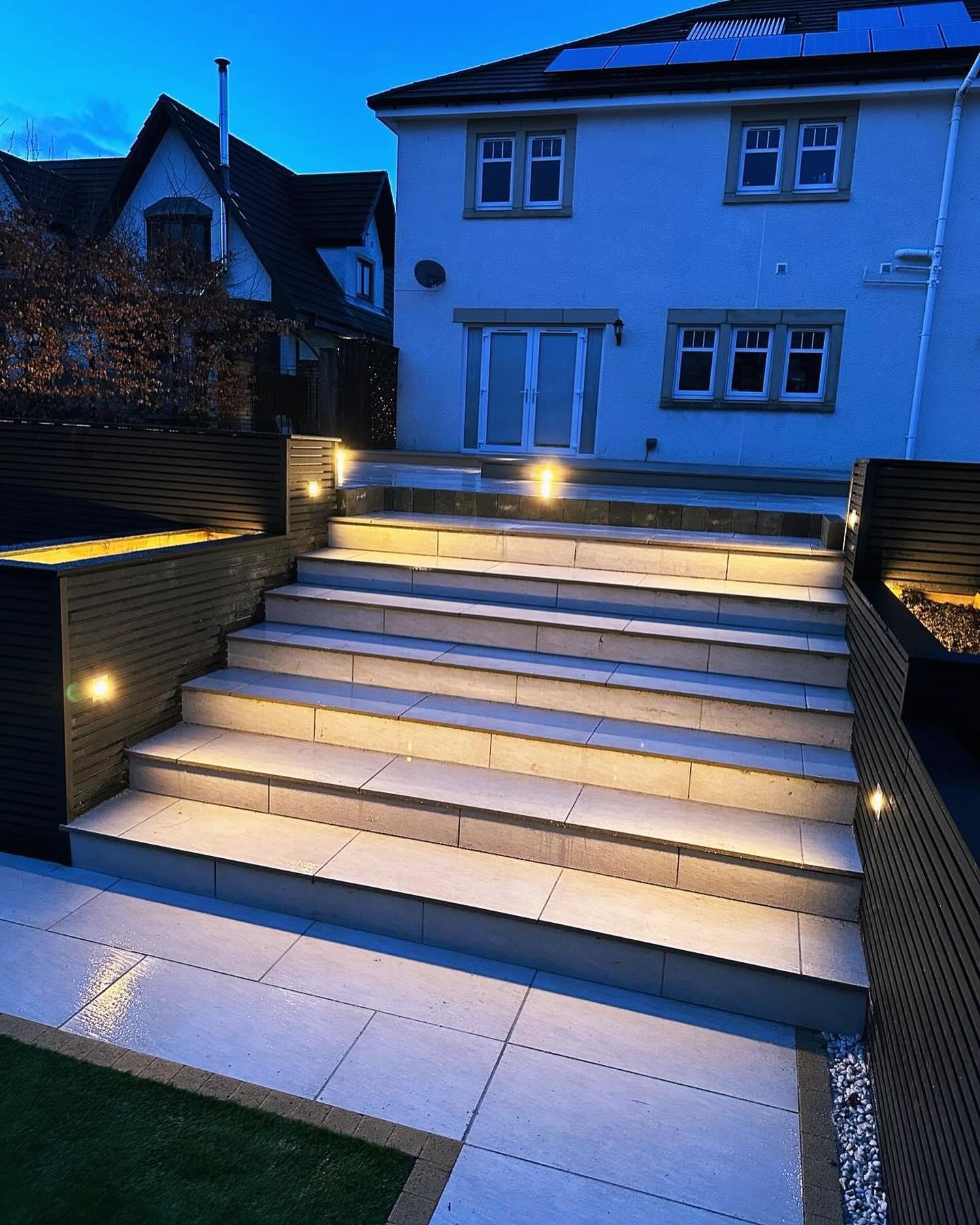 Wide porcelain steps lit from beneath each tread, rising between charcoal-clad walls to the back of a house at blue hour.