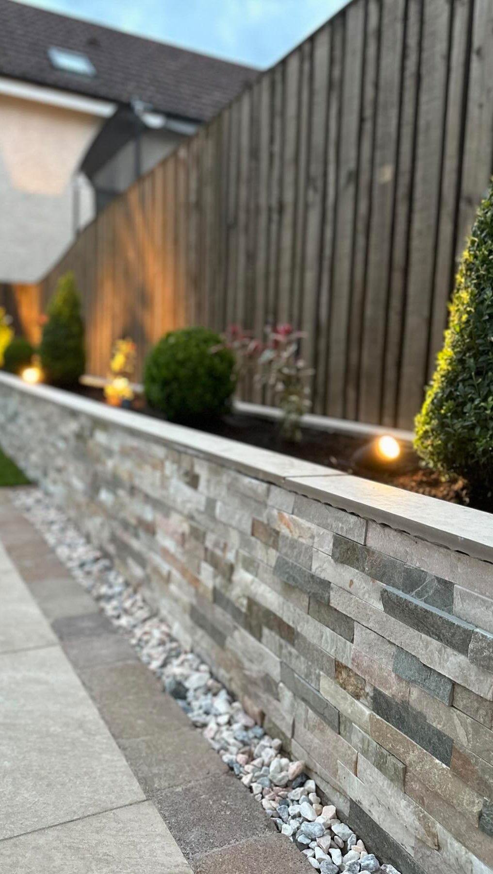 A raised planter faced in stacked stone cladding, edged with white pebbles and lit by small uplighters, with box balls and conifers along a timber fence.