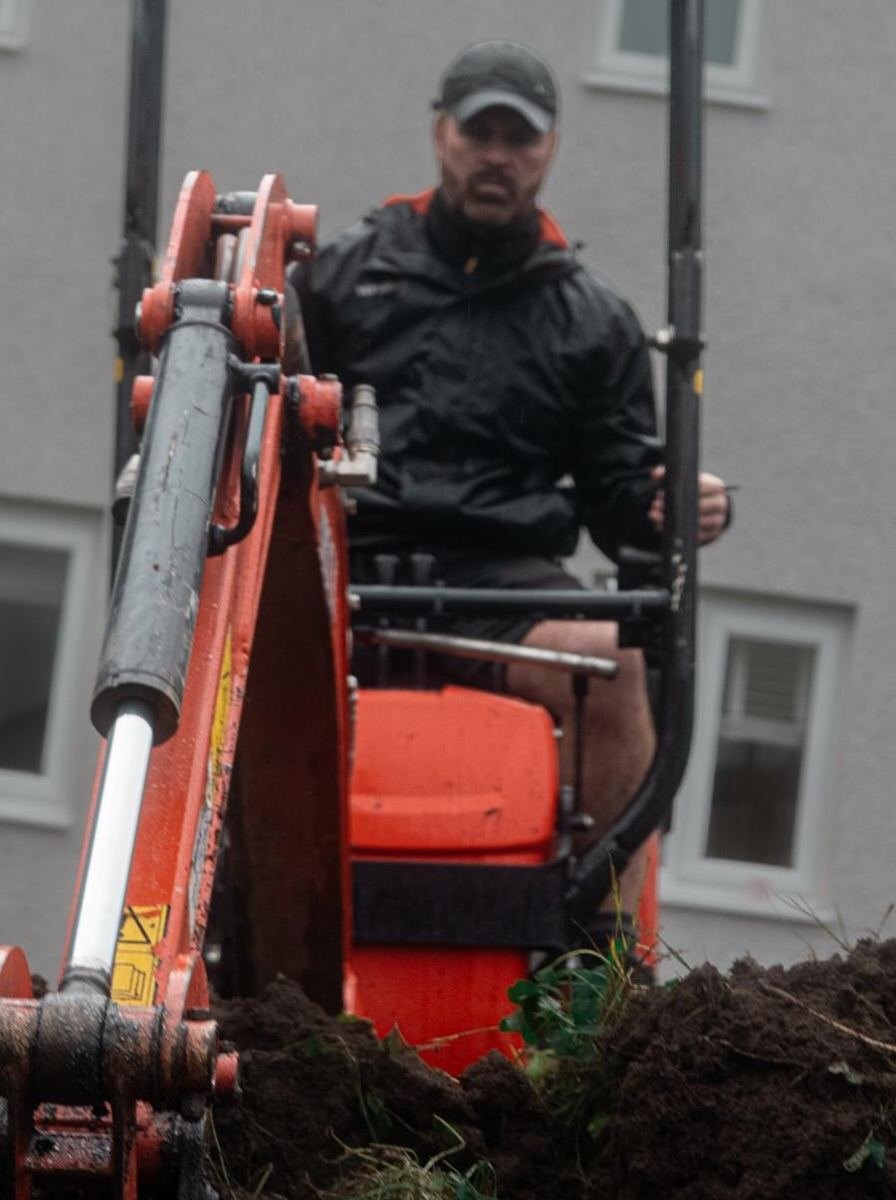 A Cornerstone team member at the controls of an orange mini digger, excavating a garden.