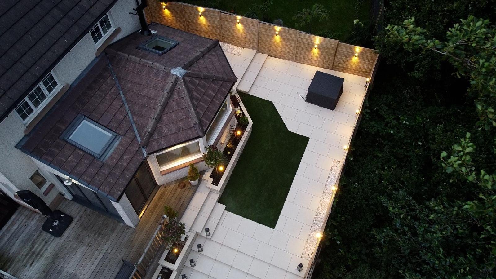 Overhead drone view of a finished garden at dusk, showing the lines of the paving, lawns, raised decking and lit planters.