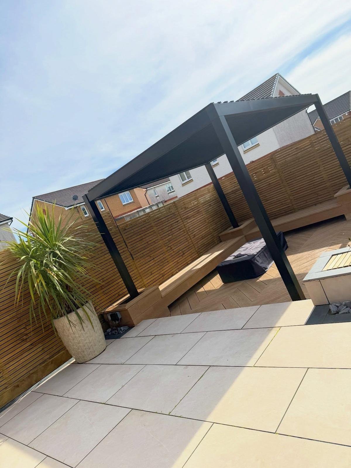 A black pergola over a seating area on pale porcelain, with a wooden hammock stand, palms in pots and slat fencing.
