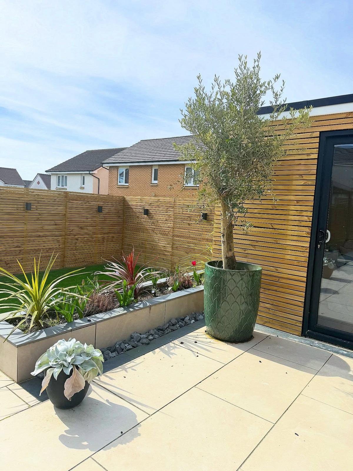 Raised planters and an olive tree in a large green pot beside the house, on a pale porcelain patio.