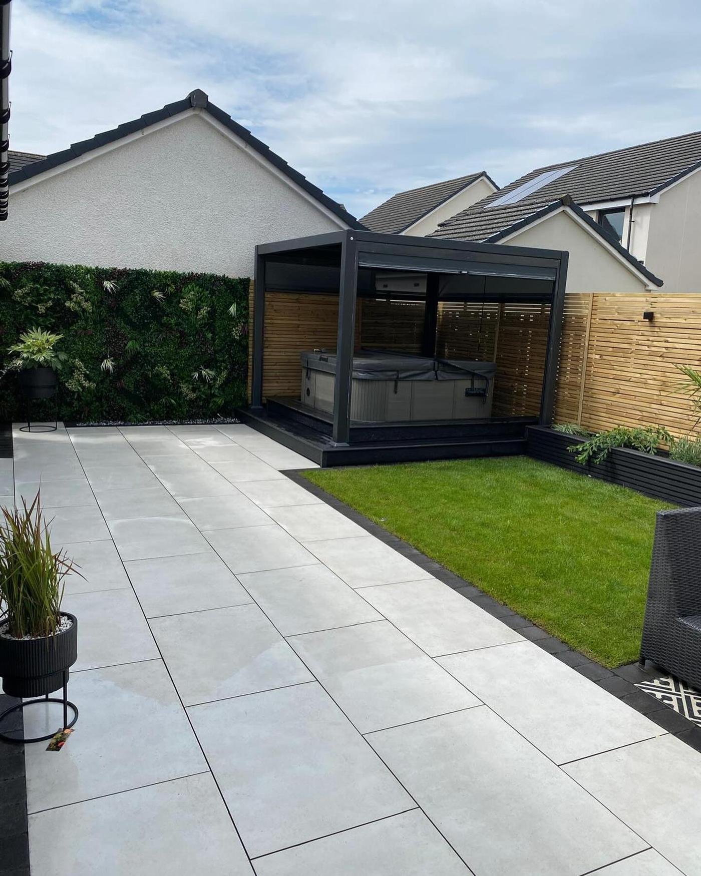 A dark aluminium pergola sheltering a hot tub at the end of a white porcelain patio, with a green foliage wall and slat fencing.