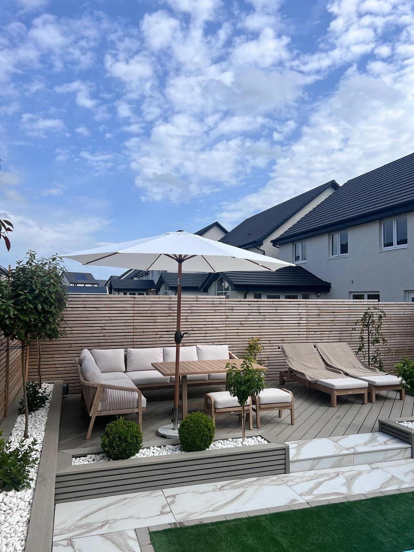 A new-build garden in neutral tones: grey composite decking with beige outdoor sofas under a parasol, horizontal slat fencing, box balls and small trees in white stones, and marble-effect porcelain.